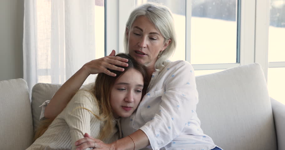 Loving mother cuddles, strokes her frustrated teenage daughter sit together on sofa. Upset adolescent 12s girl receive psychological support from mom, parent express love, hugging, consoling her child | Shutterstock HD Video #1099588985