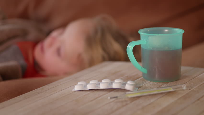 Blurred view of sick child lying on the sofa, hot drink, pills and thermometer. Ill blond girl feeling weak and tired because of fever, corona symptoms, virus disease. Healthcare concept. HQ 4K | Shutterstock HD Video #1099590571