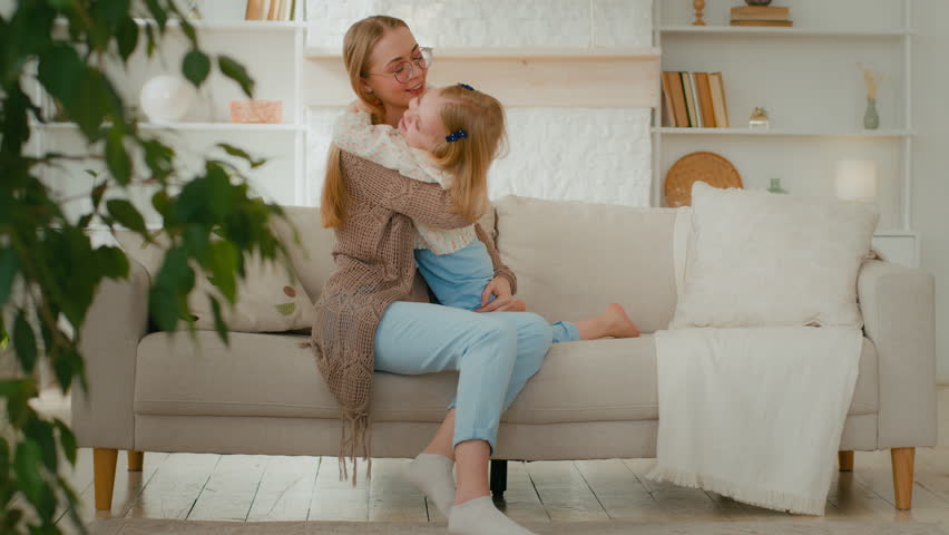 Caucasian loving mother hug cuddle embrace little daughter tickles put child down and raise up playing game play having fun with kid girl hugging embracing cuddling laughing together sit on couch sofa | Shutterstock HD Video #1099590787