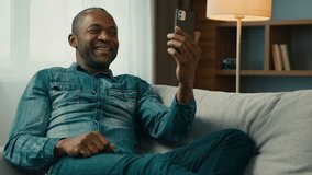 Smiling mature african american man sitting on comfortable sofa in room talk video call using mobile phone waving greet positive male remote communicate with friend by online conference on smartphone