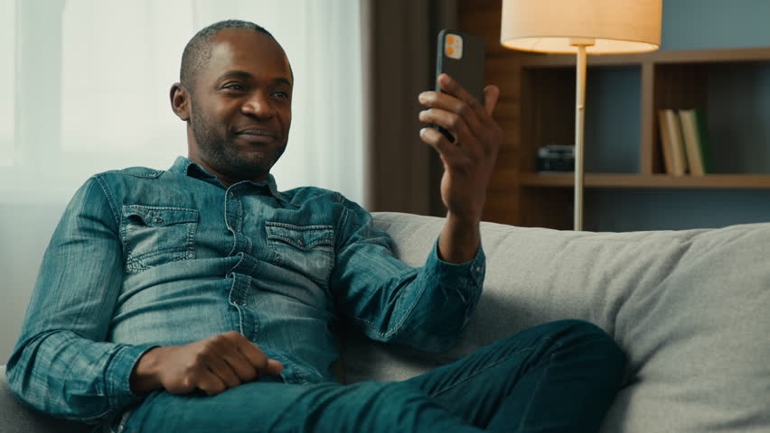 Smiling mature african american man sitting on comfortable sofa in room talk video call using mobile phone waving greet positive male remote communicate with friend by online conference on smartphone | Shutterstock HD Video #1099590789