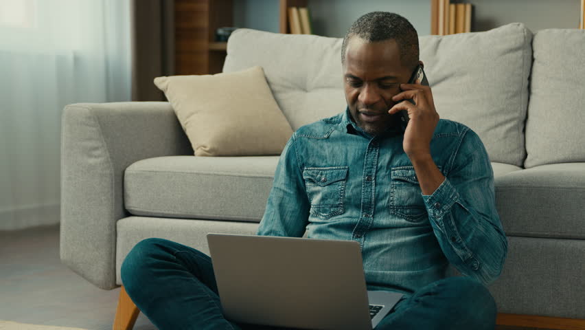 Mature african american businessman freelancer sitting in room floor talking mobile phone working on laptop ethnic man client remote make order in online store using cellphone and computer application | Shutterstock HD Video #1099590793