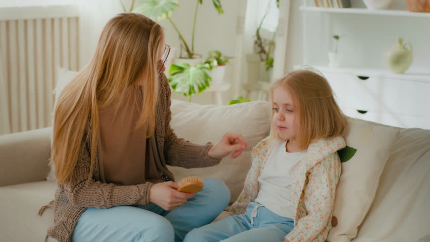 Caucasian mother remove straighten hair from face of cute child daughter after combing brushing. Mom hold comb talking conversation talk with little funny kid baby girl sitting on sofa couch at home | Shutterstock HD Video #1099590795