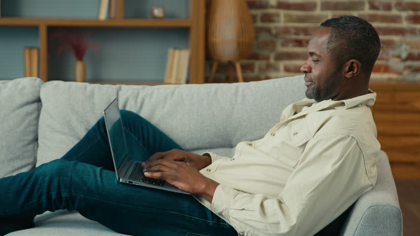 Mature african american businessman freelancer typing on laptop working on Internet lying on sofa in living room ethnic man middle-aged resting at home browsing social network checking email side view | Shutterstock HD Video #1099590805