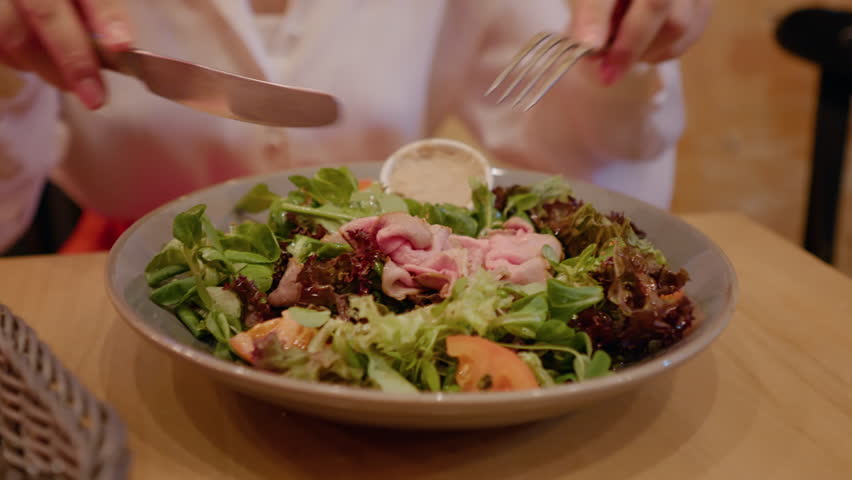 Client mixing salad using a knife and a fork while eating out. Green salad with tomatoes and ham eaten by woman.