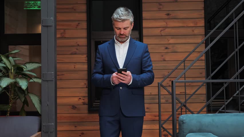 Serious mature businessman holding smartphone standing in office. Middle aged manager ceo using cell phone mobile apps Digital technology applications and solutions for business corporate development. | Shutterstock HD Video #1099596705