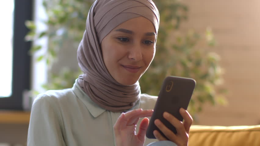 Domestic leisure. Young happy muslim lady wearing traditional headscarf reading news on smartphone online, web surfing in social media, resting on weekend at home, tracking shot, free space Royalty-Free Stock Footage #1099598525