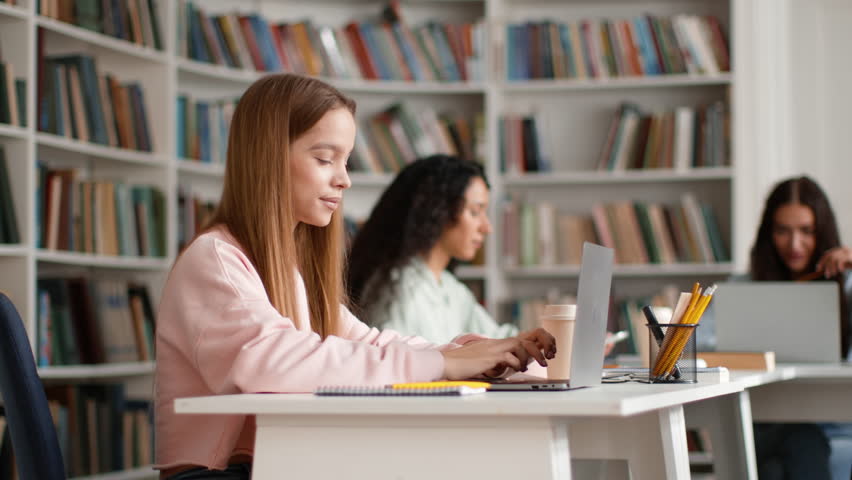 Great modern education. Young pretty smart woman student typing on laptop, turning face to camera and gesturing thumb up, studying at campus library, tracking shot, slow motion, empty space