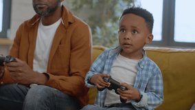 Father and son entertainment. Cute little african american boy playing video games with joystick at home, competing with his emotional daddy in car race, tracking shot, slow motion, free space