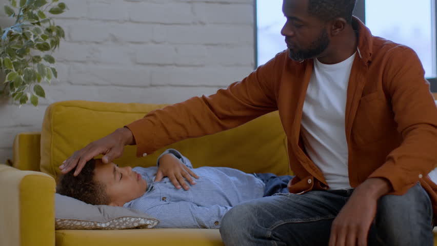 Seasonal virus. Cute little sick african american boy napping on sofa, his caring father touching forehead and cheeks, checking body temperature, tracking shot, slow motion, empty space | Shutterstock HD Video #1099598561
