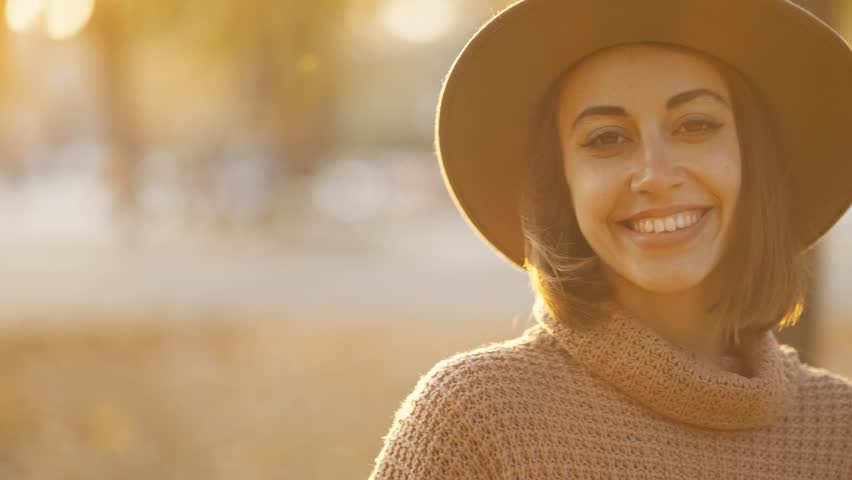 Close up autumn portrait of beautiful smiling woman wearing in wrown dress, sweater and hat. girl holds yellow leaves and playfuly looking at camera | Shutterstock HD Video #1099599031