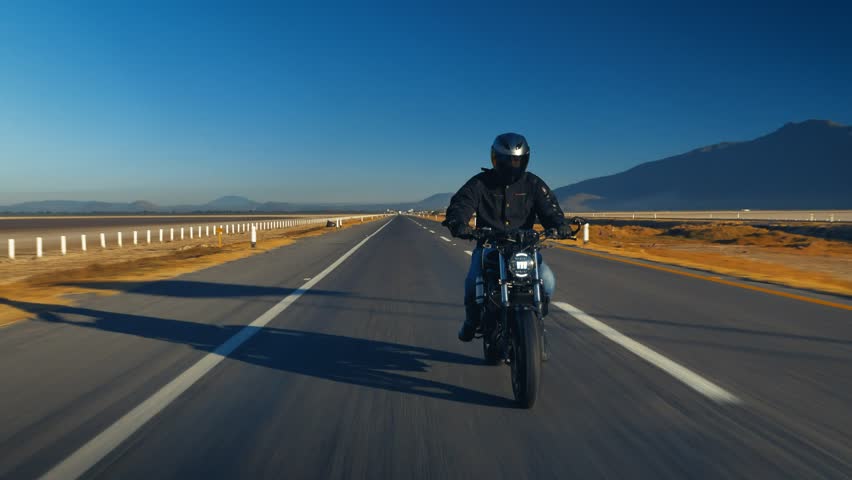 A magnificent sight of a lone man riding a motorcycle on a sunny day along a winding, empty road. The bright sun highlights the backdrop of the majestic landscape, encompassing the beauty and serenity Royalty-Free Stock Footage #1099599571