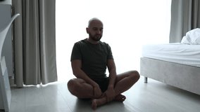 Relaxed, calm, confident bald unshaven man sitting in yoga lotus pose with crossed legs near bed in light bedroom. 