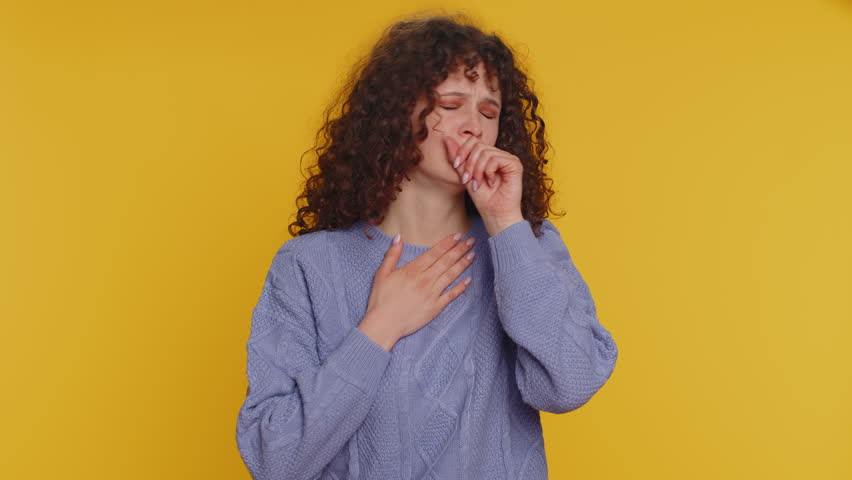 Unhealthy curly haired woman coughing covering mouth with hand, feeling sick, allergy, viral infection symptoms, coronavirus pandemic. Young adult teen girl isolated alone on yellow studio background | Shutterstock HD Video #1099602907