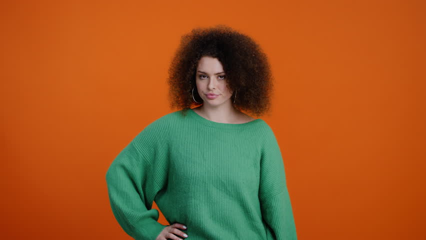 Caucasian young curly-haired brunette woman in warm green sweater, kicking her palm by her first, gesturing, showing threat sign with hands, looking at camera, standing isolated on orange background