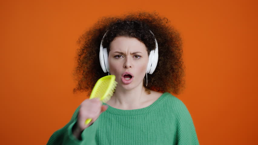 Caucasian attractive curly-haired attractive woman listening to a cool track, enjoying melody and singing into a yellow hairbrush as if into a microphone, dancing and moving isolated orange background