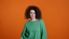 Slow motion video with a Caucasian charming attractive brunette woman, with stylish afro hairstyle, wearing warm knitted green sweater, having fun, dancing, smiling over a color orange background