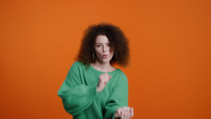 Slow motion video with a Caucasian charming attractive brunette woman, with stylish afro hairstyle, wearing warm knitted green sweater, having fun, dancing, smiling over a color orange background Royalty-Free Stock Footage #1099603349