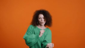 Slow motion video with a Caucasian charming attractive brunette woman, with stylish afro hairstyle, wearing warm knitted green sweater, having fun, dancing, smiling over a color orange background