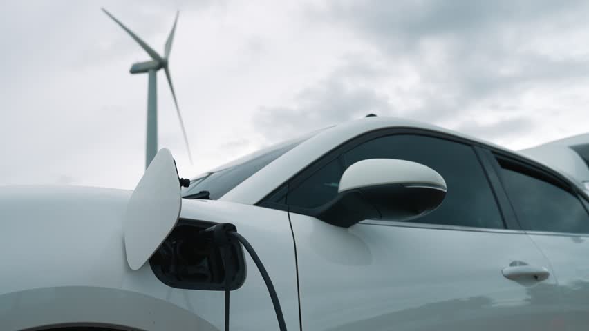 Progressive future energy infrastructure concept of electric vehicle being charged at charging station powered by green and renewable energy from a wind turbine in order to preserve the environment. | Shutterstock HD Video #1099603569
