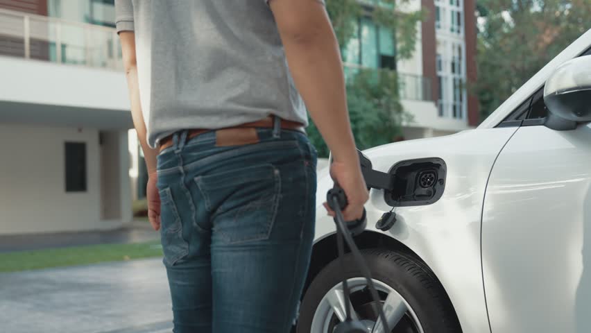 Progressive man attaches an emission-free power connector to the battery of electric vehicle at his home. Electric vehicle charging via cable from charging station to EV car battery | Shutterstock HD Video #1099603583
