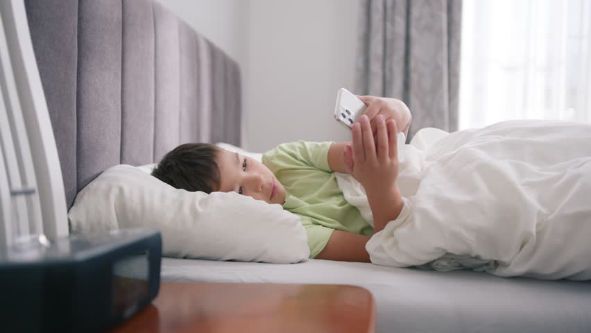 Cute mixed race kid boy holding smart phone enjoying using mobile apps, playing games at bed. Small mixed race child learning in cellphone, watching video, having fun with mobile technology concept 4K Royalty-Free Stock Footage #1099604409