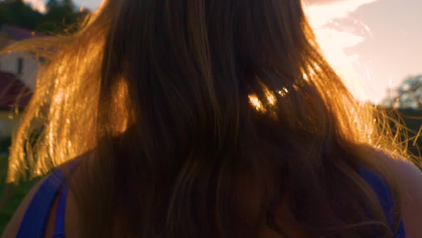 CLOSE UP, LENS FLARE: Gentle summer wind blowing through woman's long wavy hair. Rear view of beautiful young brunette, illuminated with amazing golden light, is walking towards the setting summer sun Royalty-Free Stock Footage #1099604467
