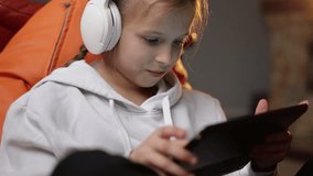 Excited teen girl plays game at home on digital tablet technology device sitting on sofa. Emotional child in headphones holds pad computer surfing internet.Children tech addiction concept.