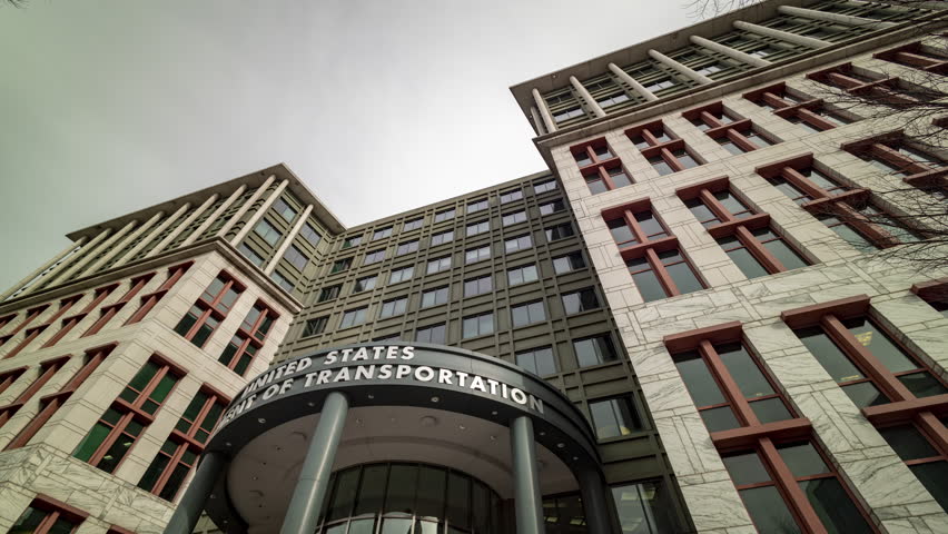 A time-lapse video shows the exterior of the US Department of Transportation building in Washington, DC, revealing the blurred motion of people coming and going during the day. The camera tilts down. Royalty-Free Stock Footage #1099607301