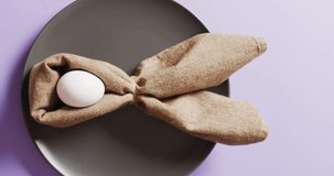 Vertical video of egg and ribbon in shape of rabbit on plate on purple background with copy space. Easter, tradition and celebration concept.