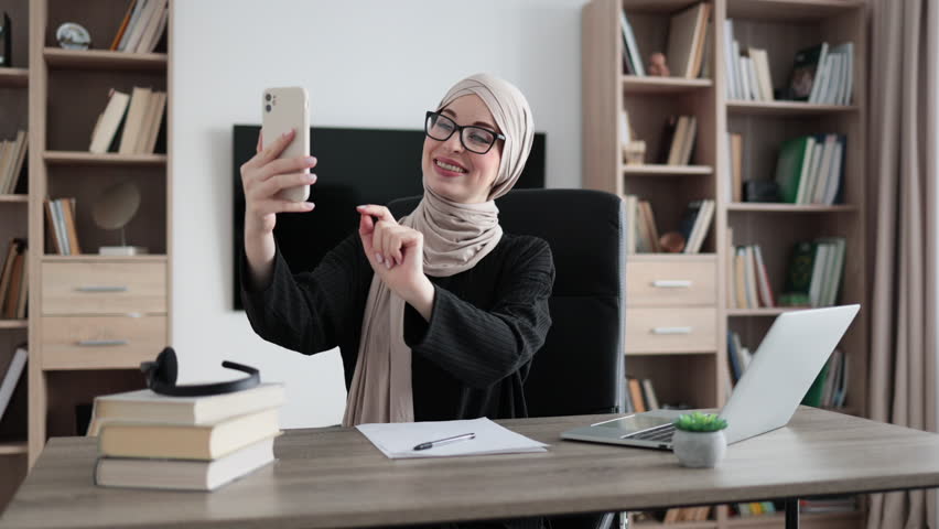 Portrait of smiling muslim businesswoman having mobile video conversation while sitting at table and working on wireless laptop. Working process at office of young girl in hijab.