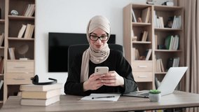 Beautiful young concentrated hardworking muslim businesswoman, it specialist or manager in hijab typing message on modern smartphone and working on laptop computer in office.