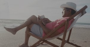 Animation of have a merry christmas over senior hispanic man sunbathing with book on beach. Christmas, family, relations and tradition concept digitally generated video.