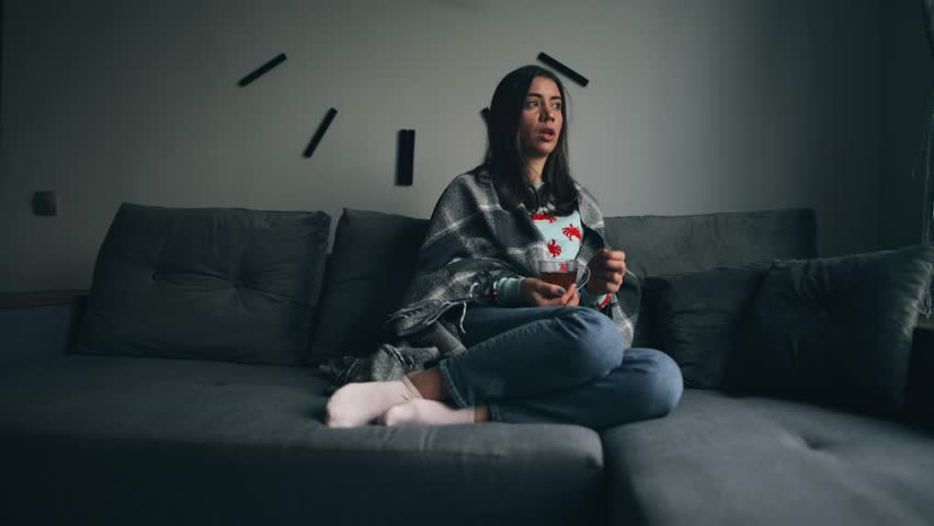 A young woman has a cold, coughs hard and drinks hot tea, sits on the sofa at home, wrapped in a blanket | Shutterstock HD Video #1099616203