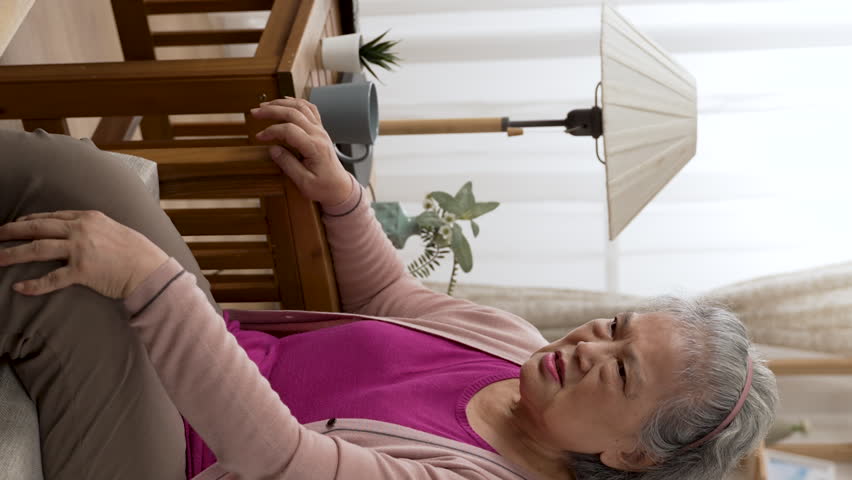 Vertical Screen: asian senior female having chronic knee pain is sitting back in sofa to massage her joints with a painful look while she is trying to stand up in the living room | Shutterstock HD Video #1099616479