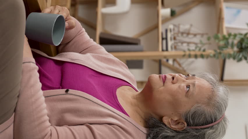 Vertical Screen: lonely asian grey haired grandma gazing into distance with an unhappy look while having tea on living room couch by herself at home | Shutterstock HD Video #1099616483