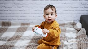 Happy cute Caucasian toddler sitting on the bed. Lovely child waves his toy phone holding in the hand. White wall backdrop.