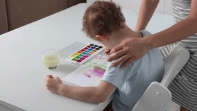 Little boy painting with paints and a brush with his mother at a white table. The boy learns to draw with watercolor paints together with a caring mother. Mom supports the child, ruffles his hair