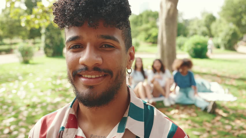 Portrait of young smiling man with curly hair wearing striped shirt posing for the camera in the park . Picnic on summer day outdoors his friends sitting in distance blurred on background