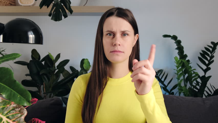Portrait of young caucasian woman warns sitting on sofa at home. Frowning serious brunette female 20s old years looking at camera and waving index finger threateningly. People emotion concept | Shutterstock HD Video #1099621011