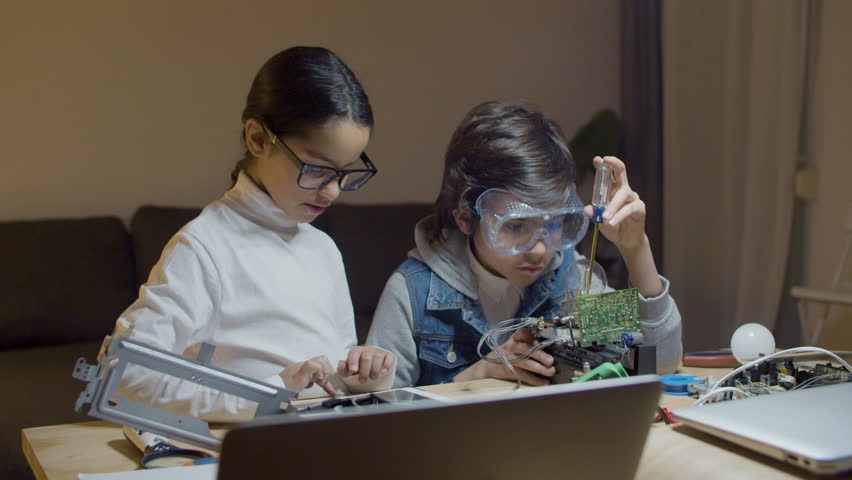 Two schoolchildren doing interesting project together. Cute girl looking at circuit on digital tablet, giving instructions while boy using tool, fixing electrical set. Education, hobby concept