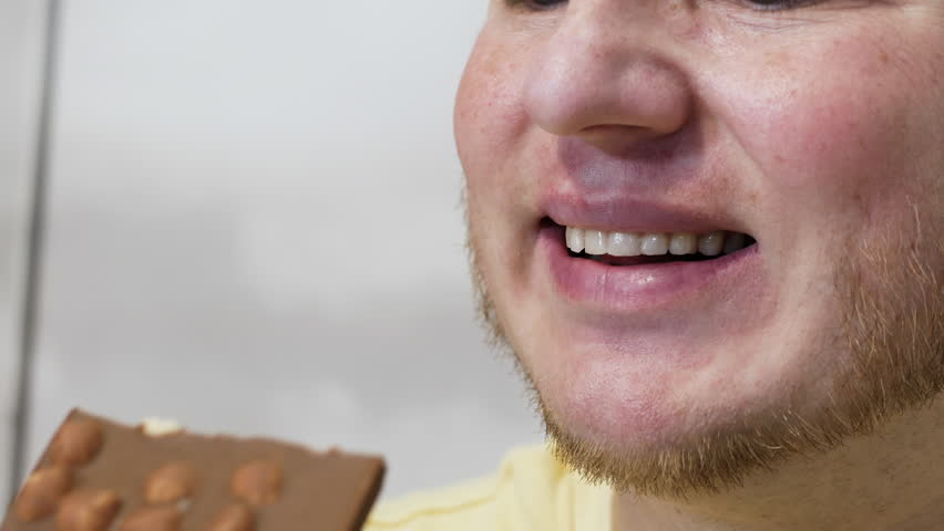 A man with a beautiful smile eats appetizing chocolate with a nut, a portrait of a man eating milk chocolate close-up. Man biting a bar of chocolate with his teeth.