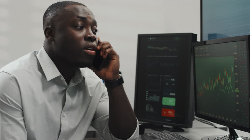 Modern young adult African American specialist looking at currency charts on computer screens working with client on phone, explaining them something