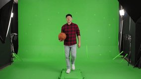 Portrait of Young Motivated Bearded Man in blue jeans, a black baseball cap and a plaid shirt, carries a basketball in his hands and walks forward against the background of a Green Screen, Chroma Key