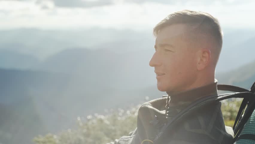 A man with a backpack takes a picture of the landscape on his phone while hiking. Portrait of a guy taking a photo with a smartphone in nature. Sunny day at the top. High quality 4k footage