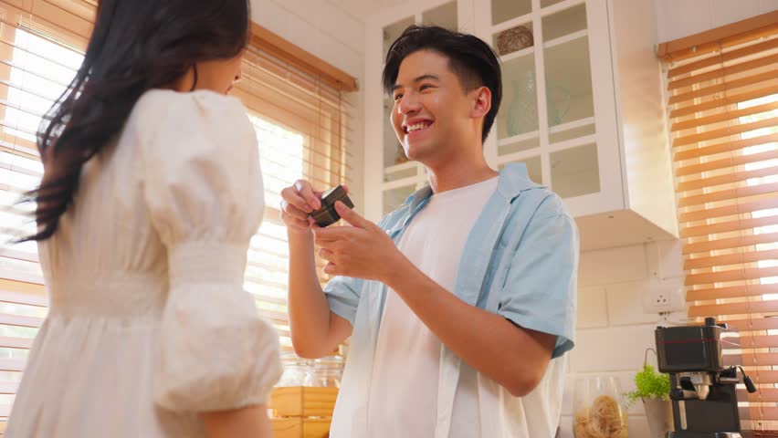 Asian romantic man making surprise proposal of marriage to girlfriend. Attractive young male and proposing to beautiful happy woman, with wedding ring enjoying surprise engagement in kitchen at home. Royalty-Free Stock Footage #1099637137