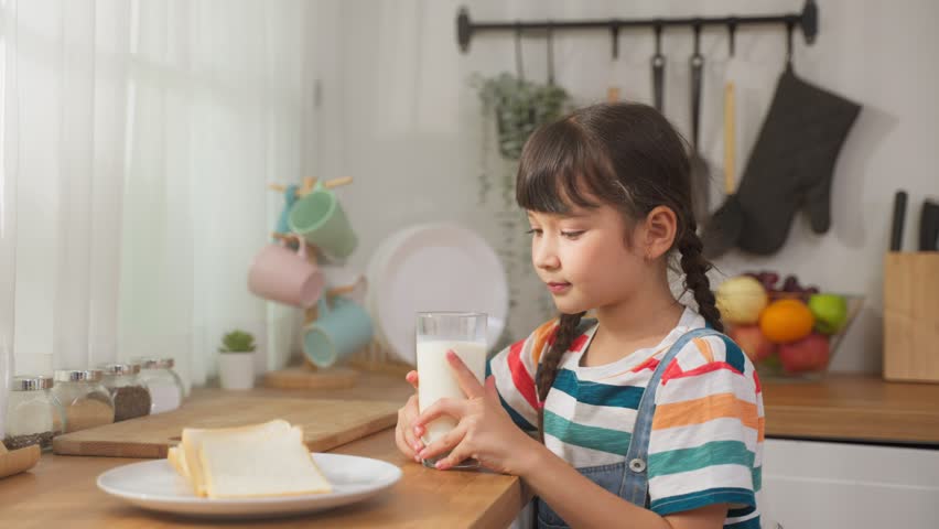 Caucasian little kid hold a cup of milk and drinking in kitchen at home. Young preschool girl or daughter sipping milk, messing up milky on moustache and lips after wake up for health care in house. Royalty-Free Stock Footage #1099637157