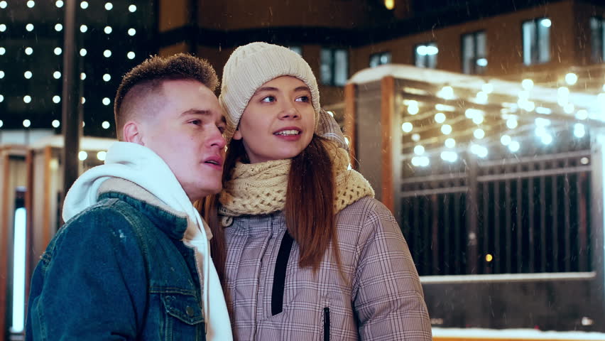 Couple girl and guy are looking at something in winter sky on evening with snowfall. Guy is showing her far away by hand. They are standing outdoors on street in the city. Romantic date, relationship.
