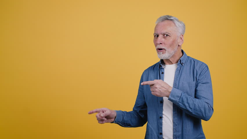 Cheerful elderly man looks to side pointing fingers and smiling. Grey-haired pensioner shows thumb up gestures standing on yellow background - Powered by Shutterstock - Get 15% off with code: PIKWIZARD15