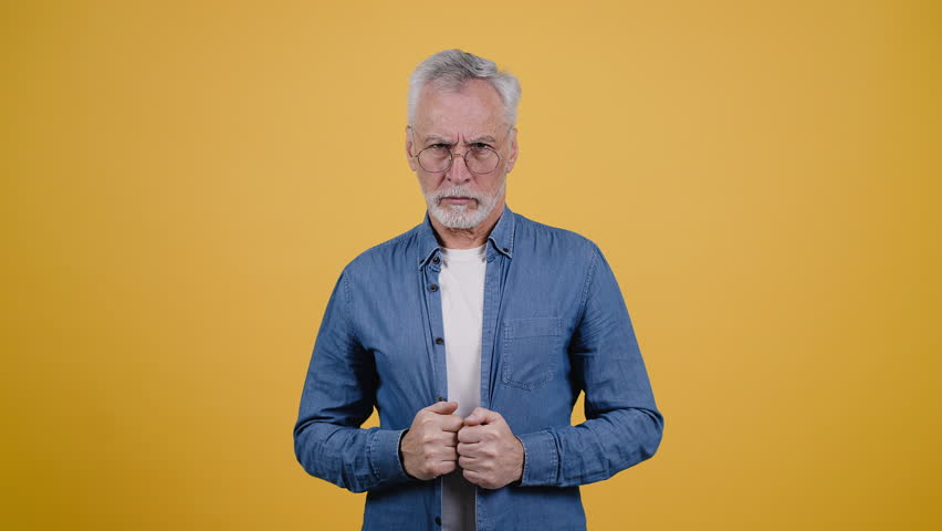 Grey-haired elderly man squints and takes off glasses. Bearded pensioner in denim shirt gets shocked and approves work achievements on yellow background